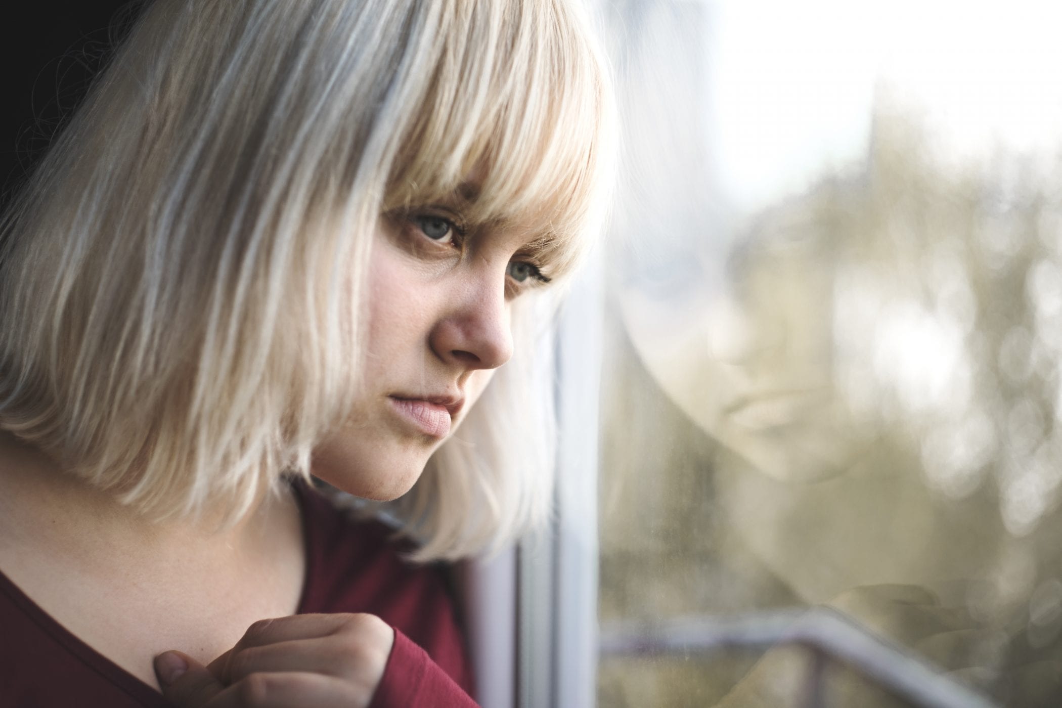 Close up of depressed young blond woman near window at home. Sadness, nostlagic, depression.